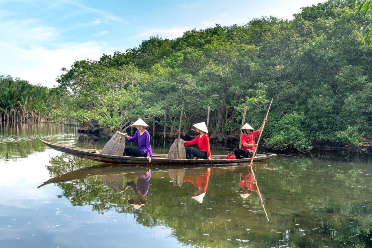 Trois femmes rament dans une barque en bois, le long d’une rive verdoyante
